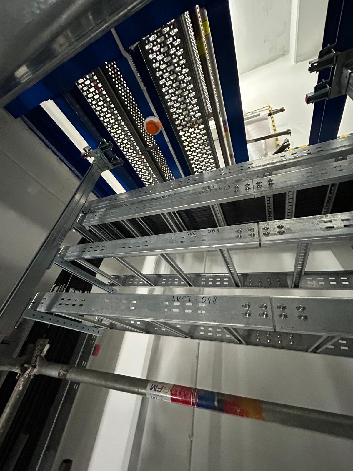 Metalowe korytka kablowe zamocowane pod sufitem, widoczne elementy rusztowania w dolnej części kadru, instalacja w trakcie realizacji.