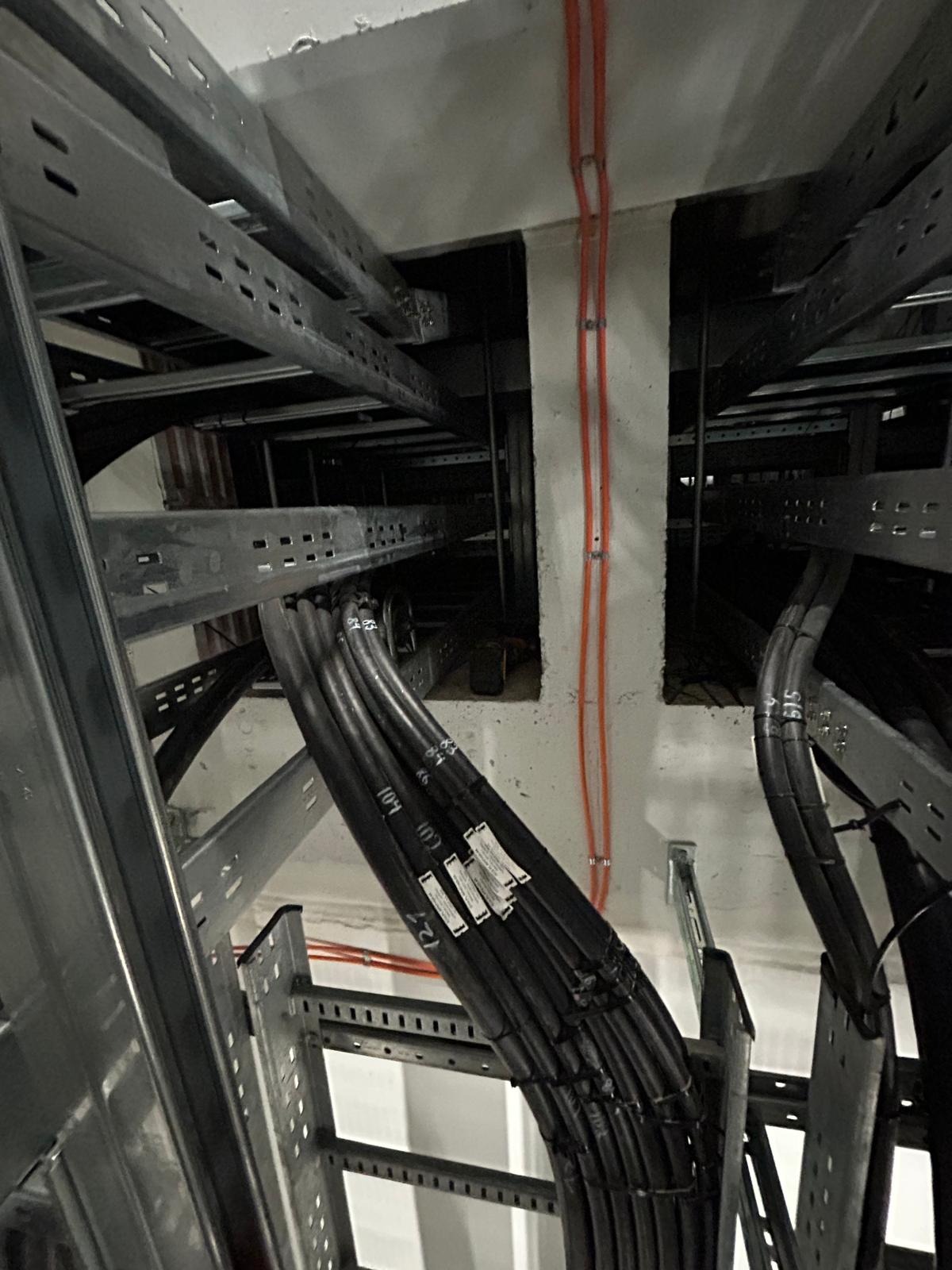Wiązka czarnych kabli elektrycznych ułożona na metalowych korytach pod sufitem, widoczne etykiety na kablach i pomarańczowe przewody na ścianie.