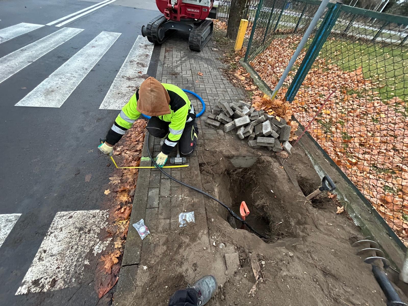 Pracownik w odblaskowej kurtce mierzy żółtą miarką odległość przy wykopie obok chodnika, w tle minikoparka i stos kostki brukowej.