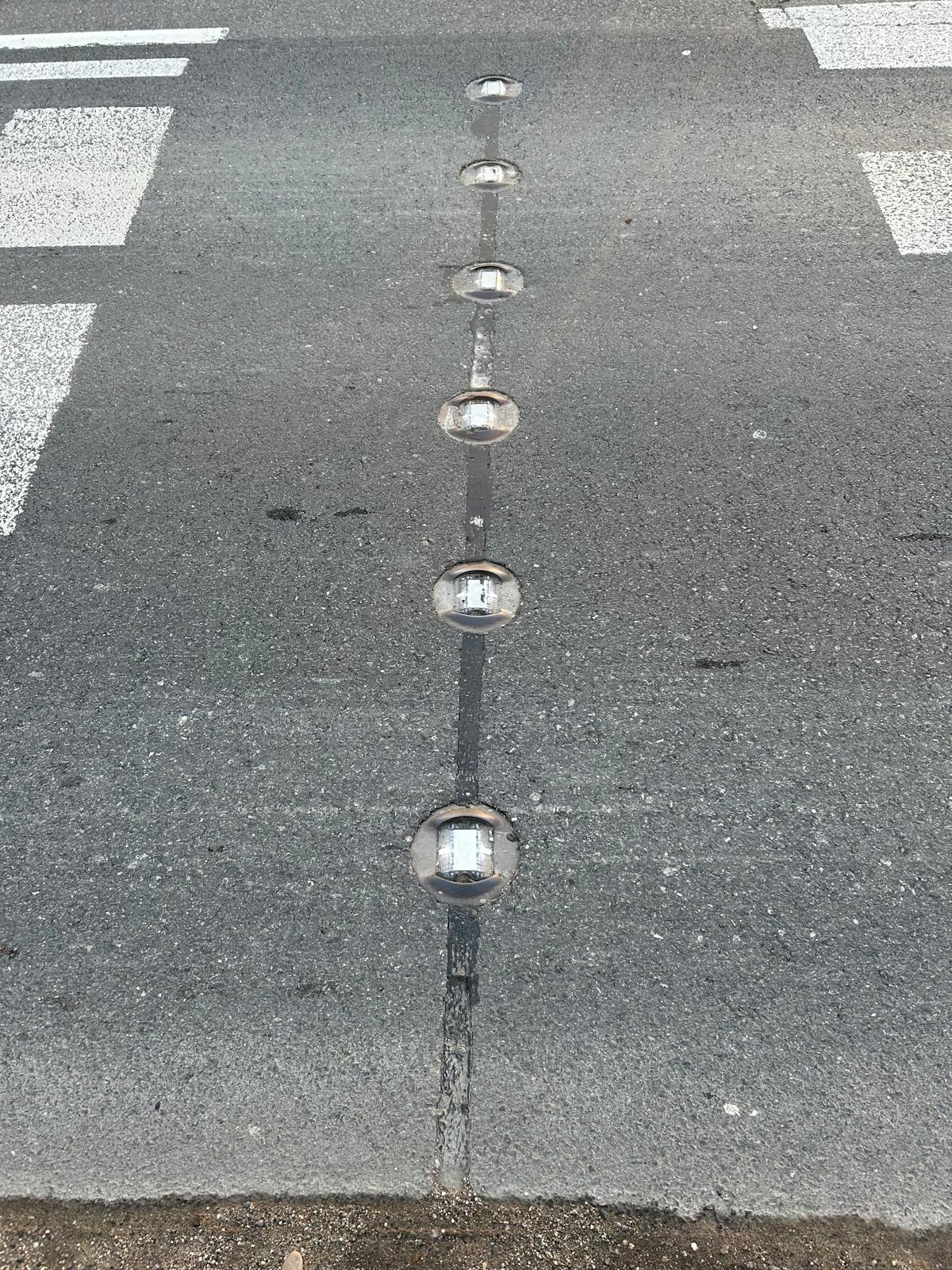 Rząd wbudowanych w asfalt, okrągłych, odblaskowych świateł LED na przejściu dla pieszych, widocznych z góry, z elementami białego oznakowania poziomego.