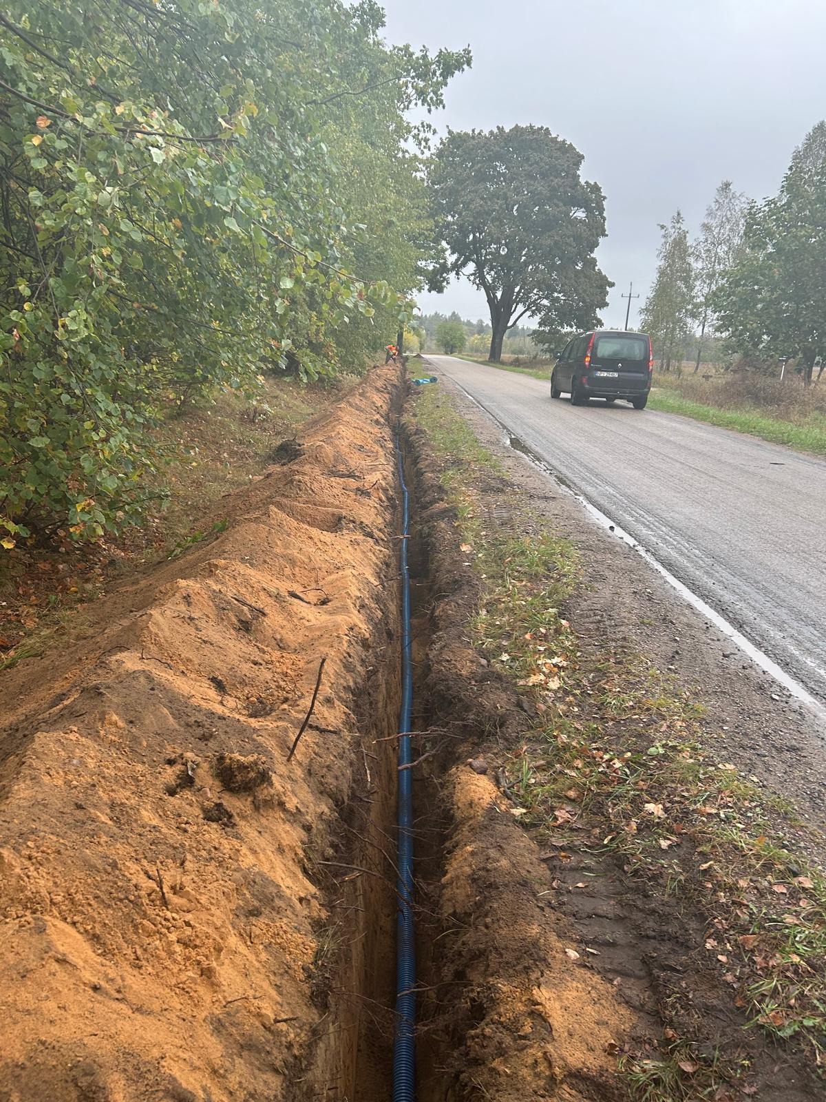 Wykop w ziemi z niebieskim kablem w peszlu, ułożonym wzdłuż drogi asfaltowej, w tle widoczny samochód i drzewa.