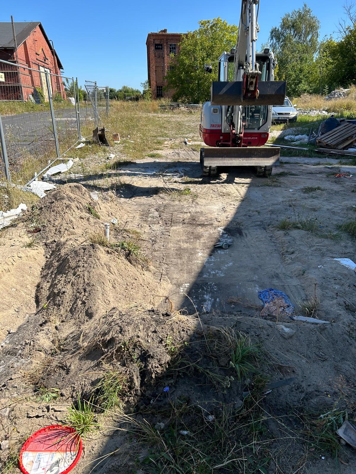 Mini koparka TAKEUCHI TB230 podczas prac ziemnych na terenie budowy, wykop pod instalację, w tle budynki z czerwonej cegły i metalowe ogrodzenie.