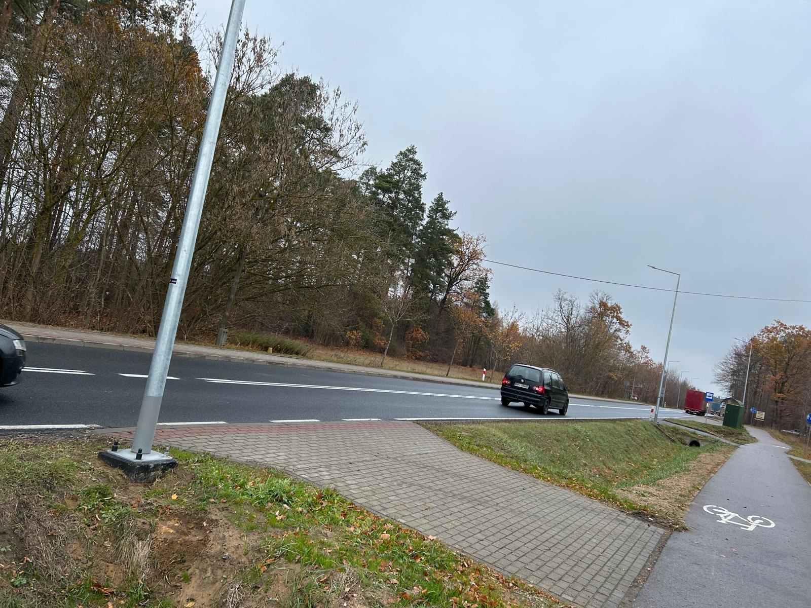 Słup oświetleniowy przy drodze, widoczne mocowanie do podłoża, w tle jesienny krajobraz i przejeżdżające samochody.