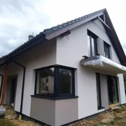 Nowoczesny dom jednorodzinny po częściowym ociepleniu, z fragmentem elewacji pomalowanym na szaro, widoczne zabezpieczenie folią balkonu przed dalszymi pracami.