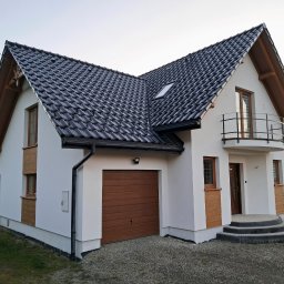 SZWED-BUD Piotr Szwed - Nowoczesny dom jednorodzinny z białą elewacją, brązowymi akcentami i ciemną dachówką. Widoczny garaż, balkon i wejście z kamiennymi schodami.