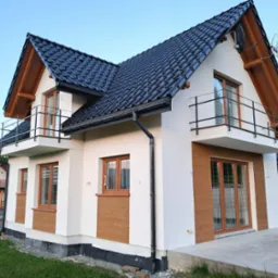 Dwukondygnacyjny dom z balkonami, białą elewacją i elementami dekoracyjnymi w kolorze drewna. Widoczna czarna dachówka i elementy orynnowania. Na dachu antena.