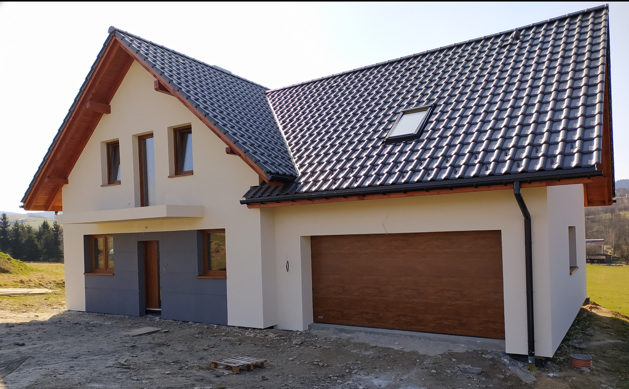 Nowo wybudowany dom jednorodzinny z jasną elewacją, grafitowym cokołem, brązową stolarką okienną i drewnianą bramą garażową, widoczne ślady prac budowlanych wokół budynku.