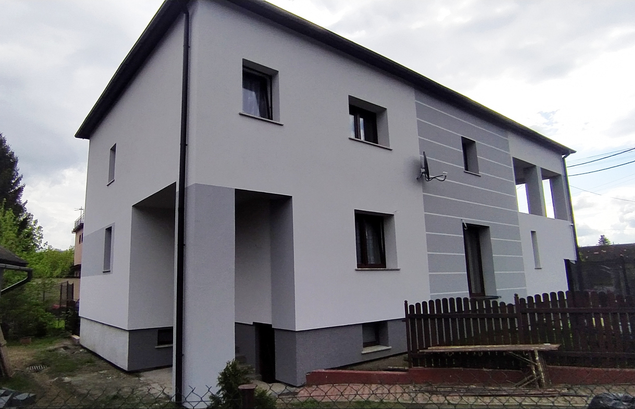 Dwukondygnacyjny dom jednorodzinny z szarą elewacją, z widocznym podziałem kolorystycznym na dolnej i górnej części fasady, z anteną satelitarną na bocznej ścianie, widziany z perspektywy ogrodu.