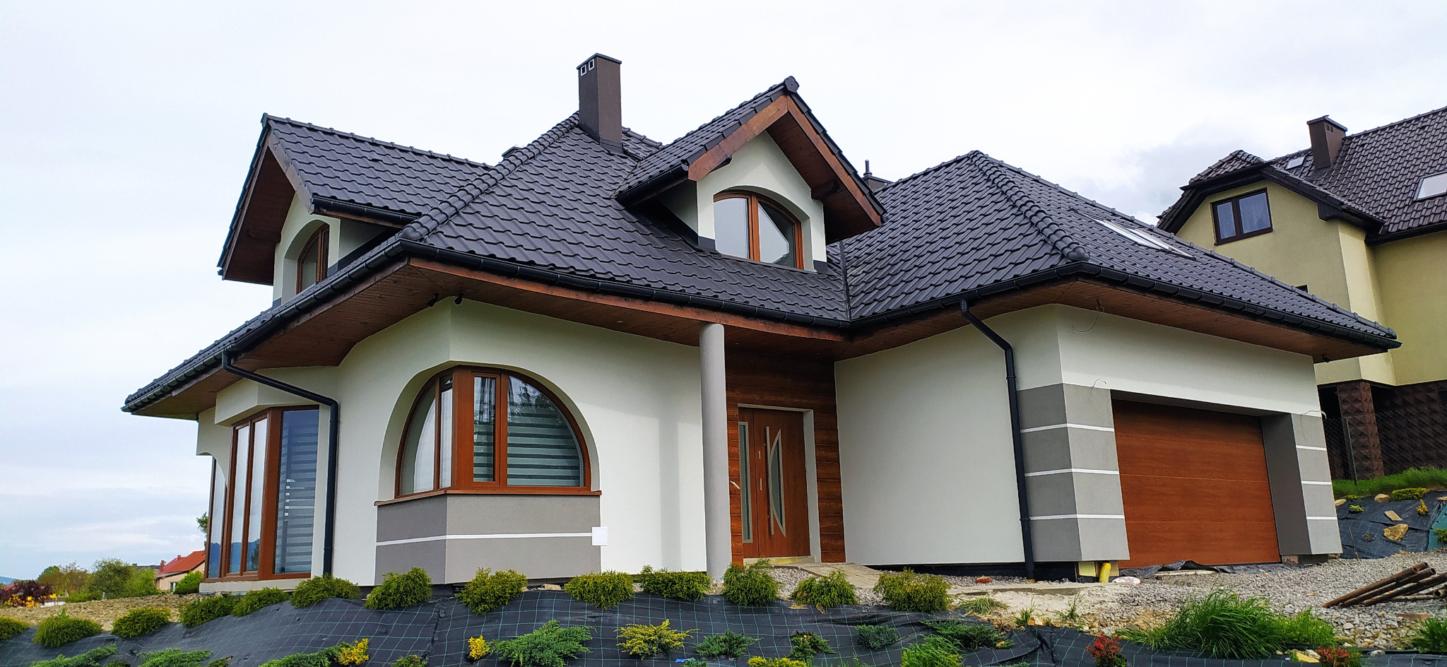 Nowoczesny dom jednorodzinny z jasną elewacją, ciemną dachówką i drewnianymi elementami wykończeniowymi, widok z poziomu ogrodu z młodymi nasadzeniami na siatce.