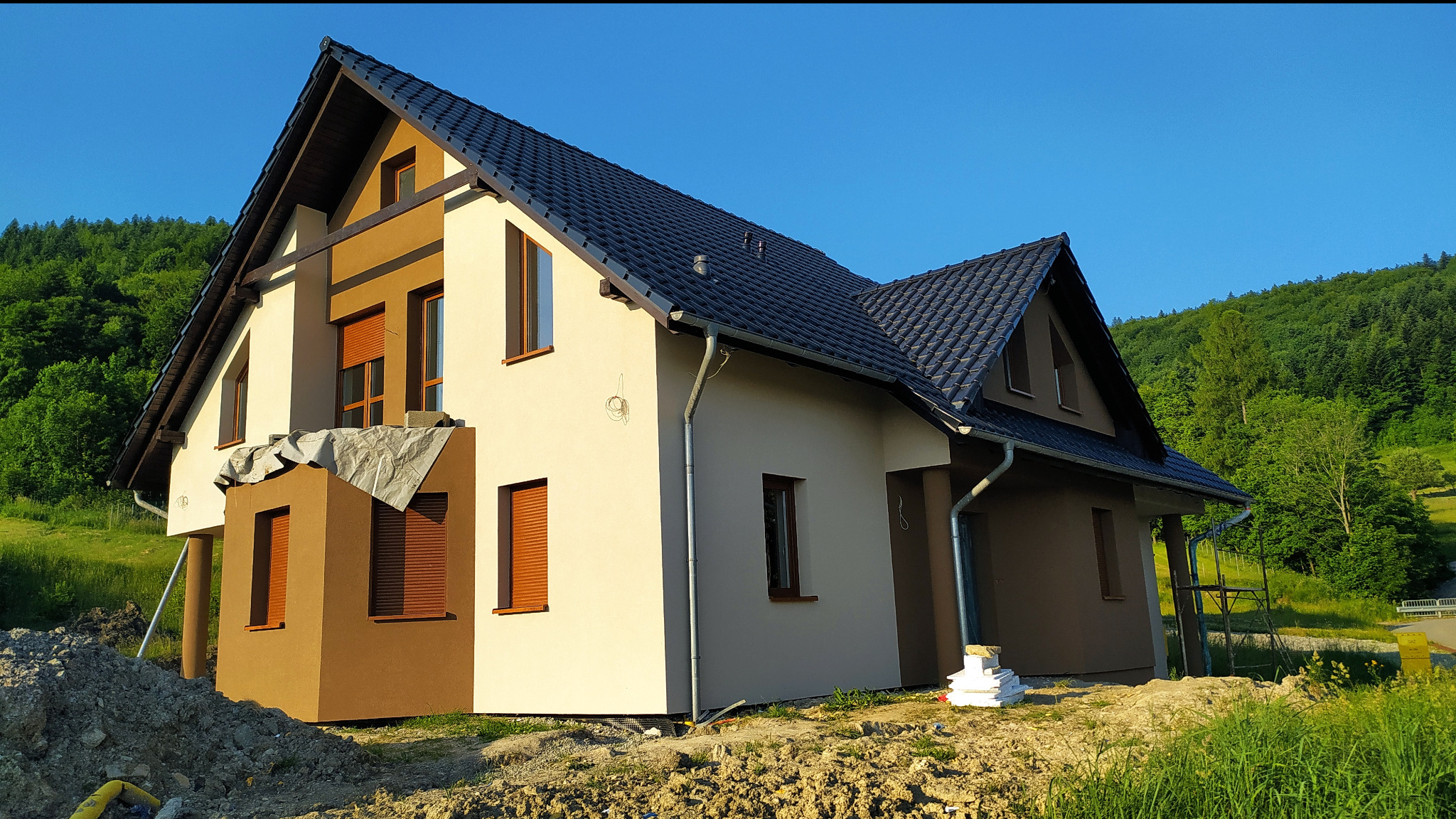 Dwukondygnacyjny dom jednorodzinny z dwuspadowym dachem, z częściowo wykończoną elewacją w odcieniach beżu i brązu, z widocznym stosem materiałów budowlanych i przygotowanym terenem wokół, na tle...