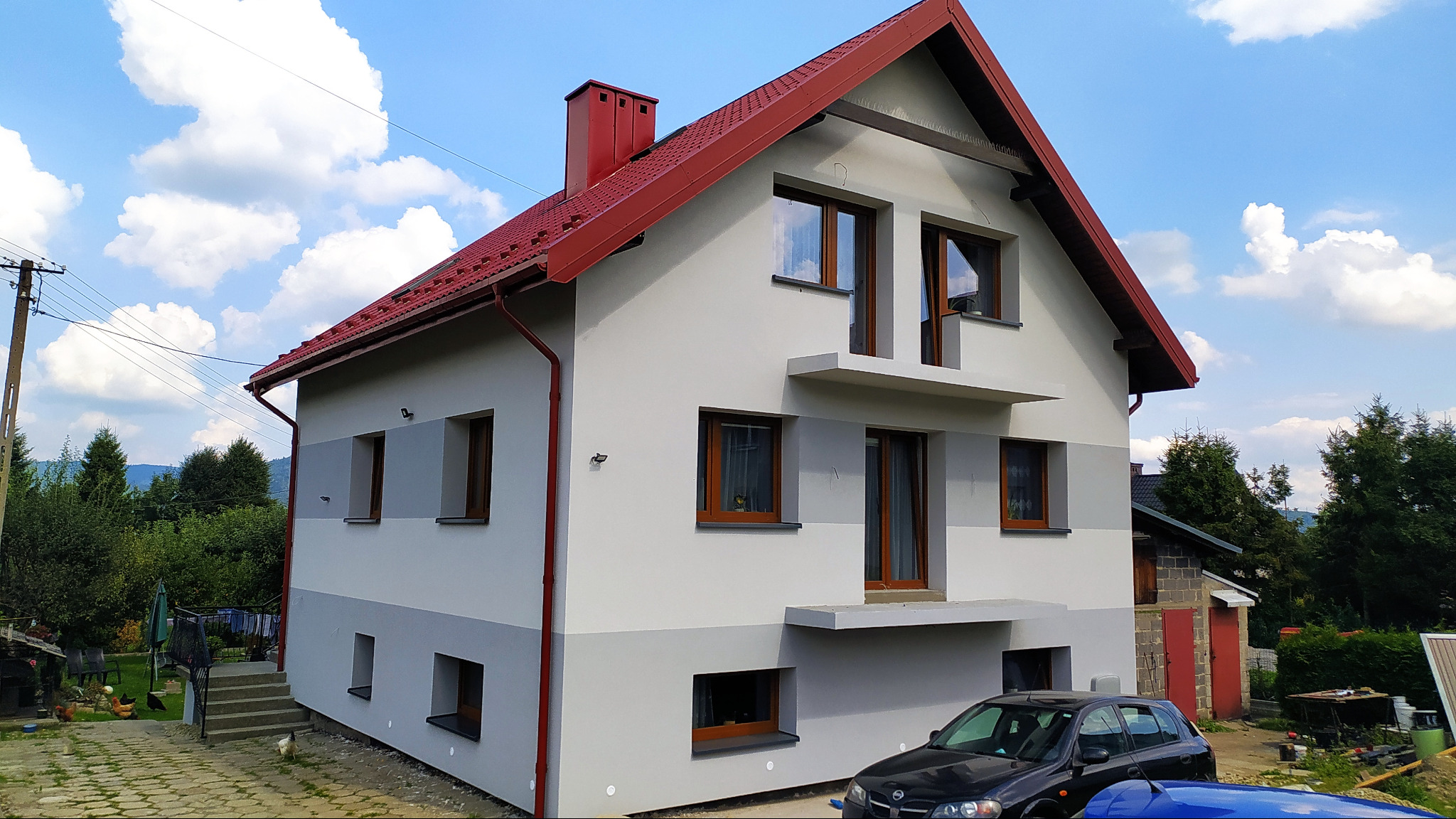 Dwukondygnacyjny dom z szarą elewacją i czerwoną dachówką, widoczny z boku na tle błękitnego nieba z obłokami. Dolna część elewacji w ciemniejszym odcieniu szarości. Na pierwszym planie zaparkowany...