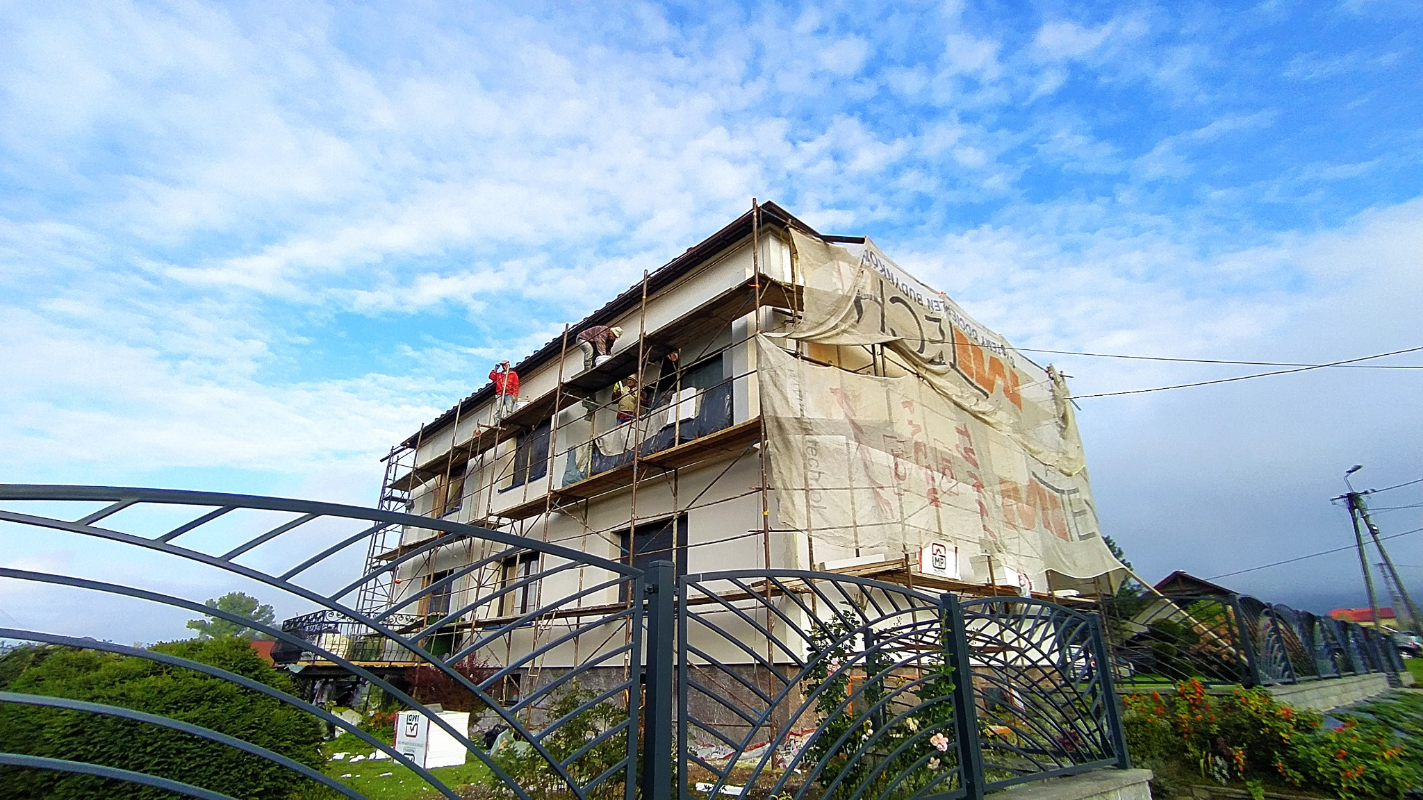 Budynek w trakcie ocieplania elewacji, widoczne rusztowania i pracownicy w strojach roboczych, siatka ochronna z nadrukiem reklamowym, ujęcie od dołu przez ozdobne metalowe ogrodzenie.