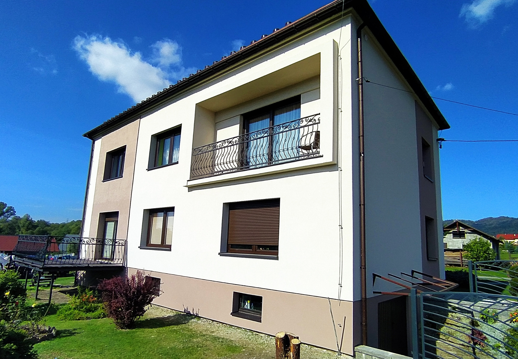 Dwukondygnacyjny dom z jasną elewacją, balkonem z ozdobną balustradą i fragmentem ogrodu, widoczne dwukolorowe wykończenie elewacji.