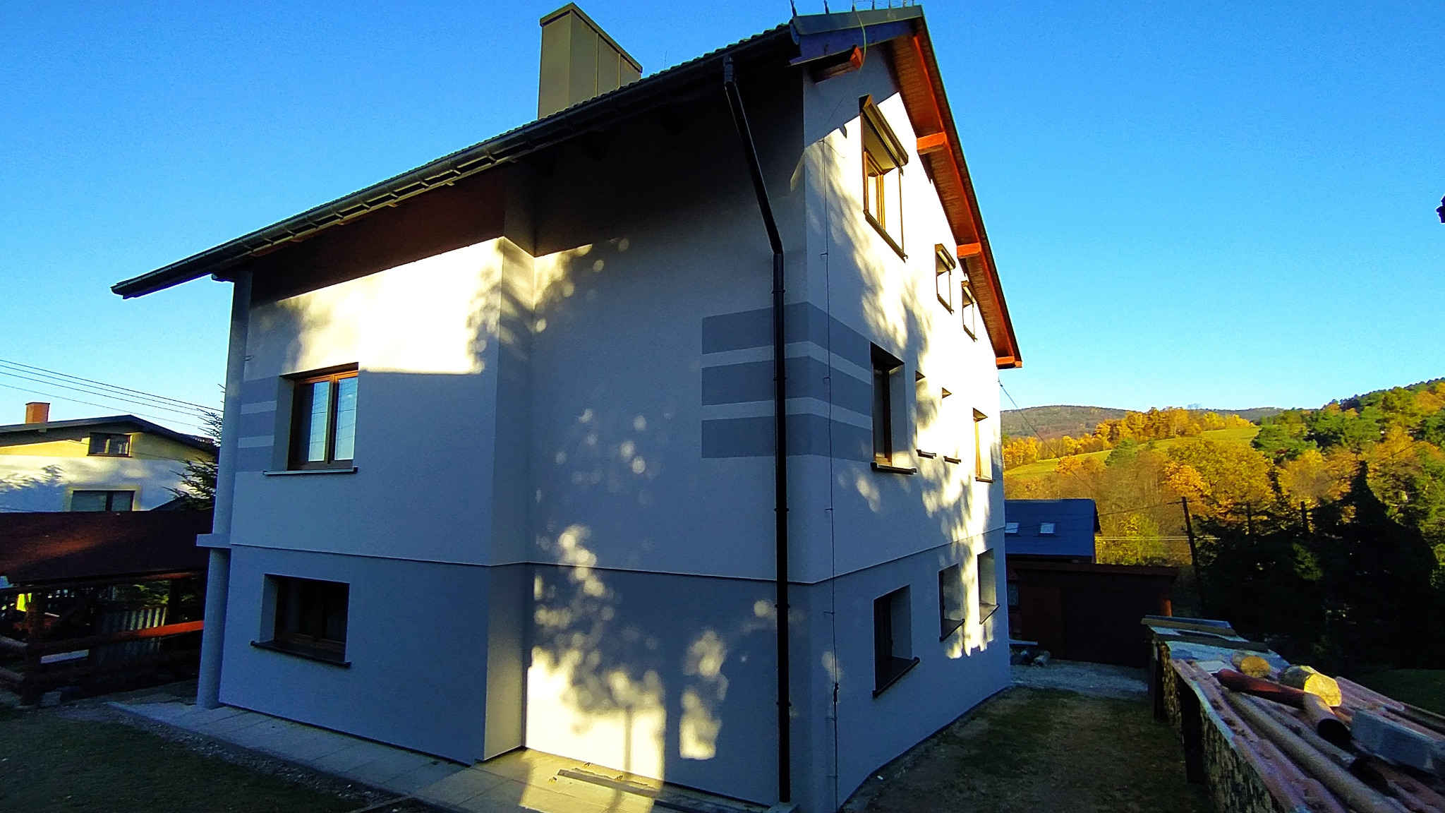 Dwukondygnacyjny dom jednorodzinny z jasnoszarą elewacją, z ciemniejszym szarym pasem u dołu i poziomymi paskami, widok z boku na tle błękitnego nieba i jesiennego krajobrazu pagórkowatego.