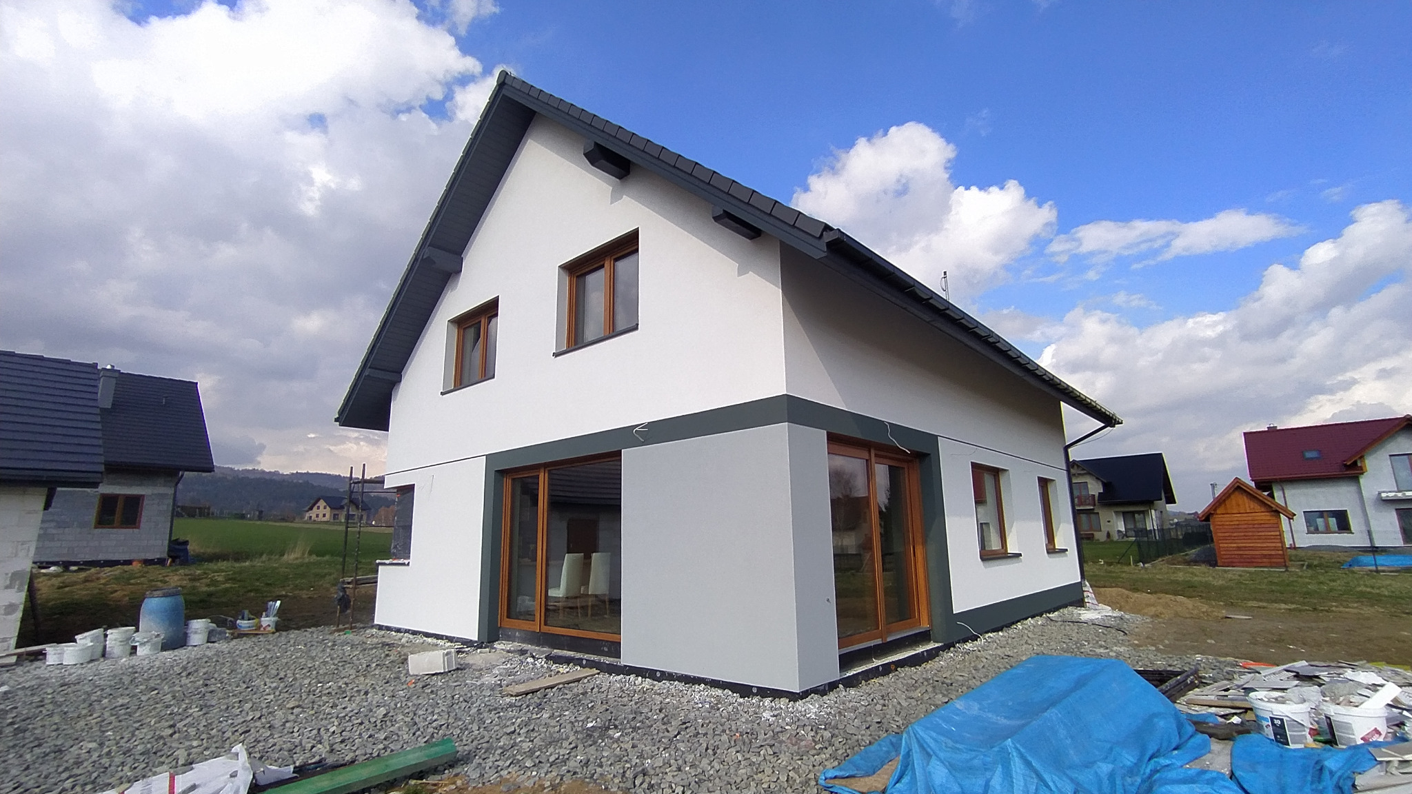 Nowoczesny dom jednorodzinny z jasną elewacją i szarymi akcentami, widok z zewnątrz, teren wokół domu w trakcie prac wykończeniowych, widoczne materiały budowlane i wiaderka z farbą.