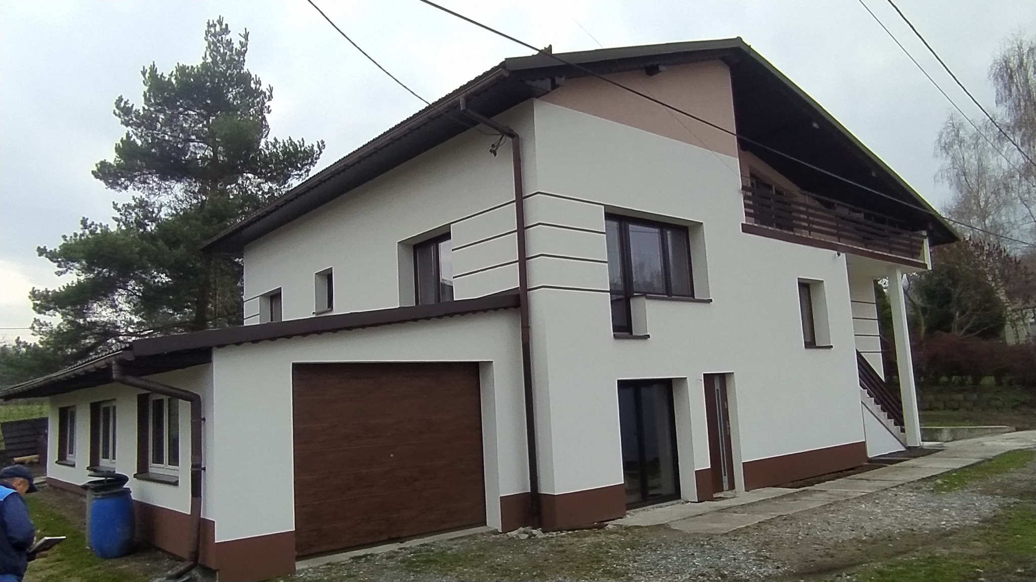 Dwukondygnacyjny dom z garażem i balkonem, pomalowany na biało z brązowymi akcentami, widok z zewnątrz na tle drzew i pochmurnego nieba.