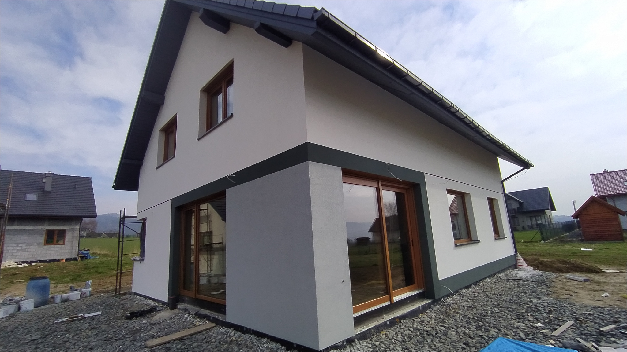 Dwukondygnacyjny dom jednorodzinny z jasną elewacją i ciemnoszarymi detalami wykończeniowymi, widoczny z boku, z elementami rusztowania i materiałami budowlanymi wokół, w tle inne budynki w trakcie...