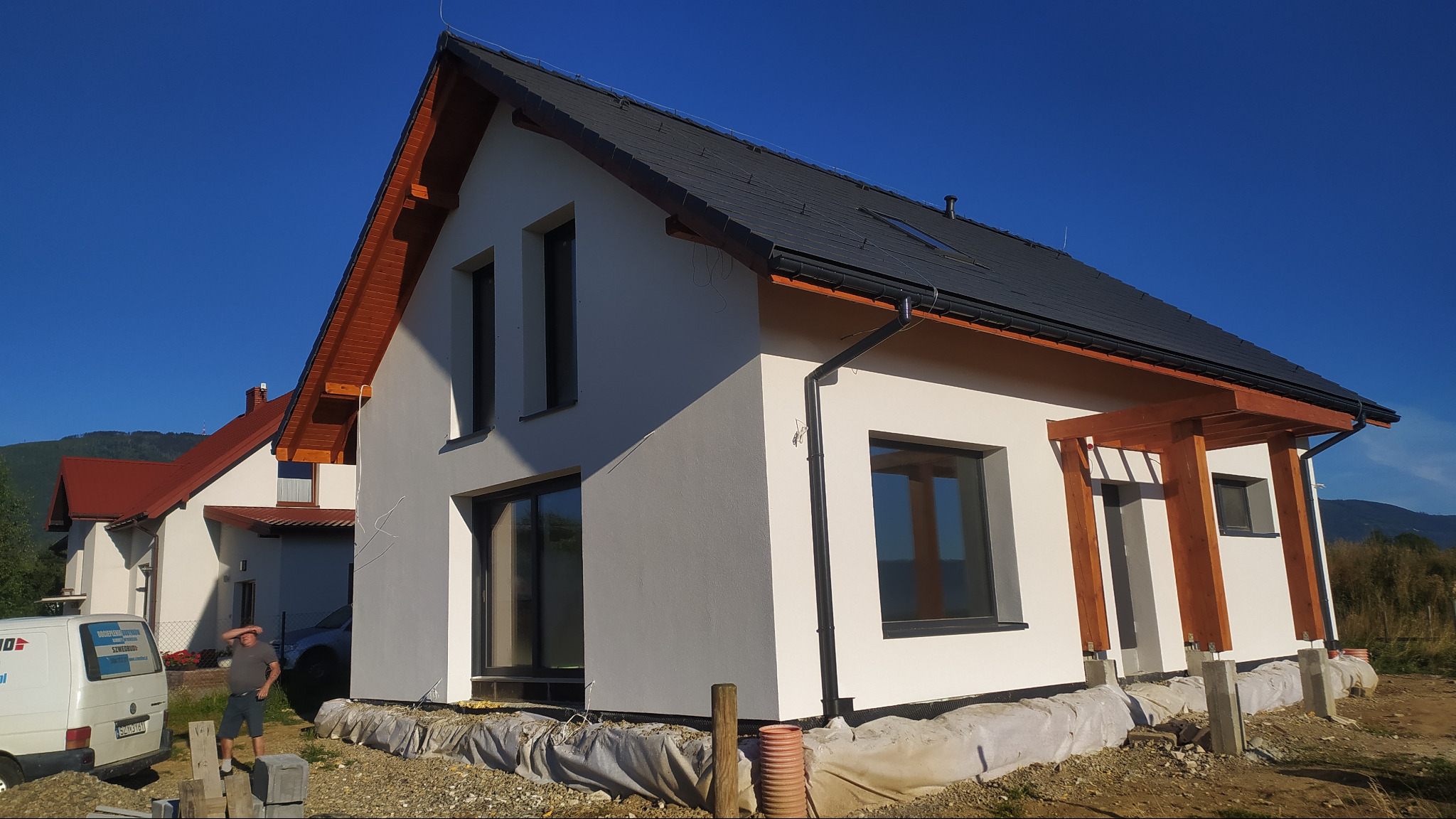 Nowoczesny dom jednorodzinny z jasną elewacją i ciemną dachówką, widoczny z boku, na pierwszym planie materiały budowlane i częściowo widoczny samochód dostawczy z reklamą firmy.