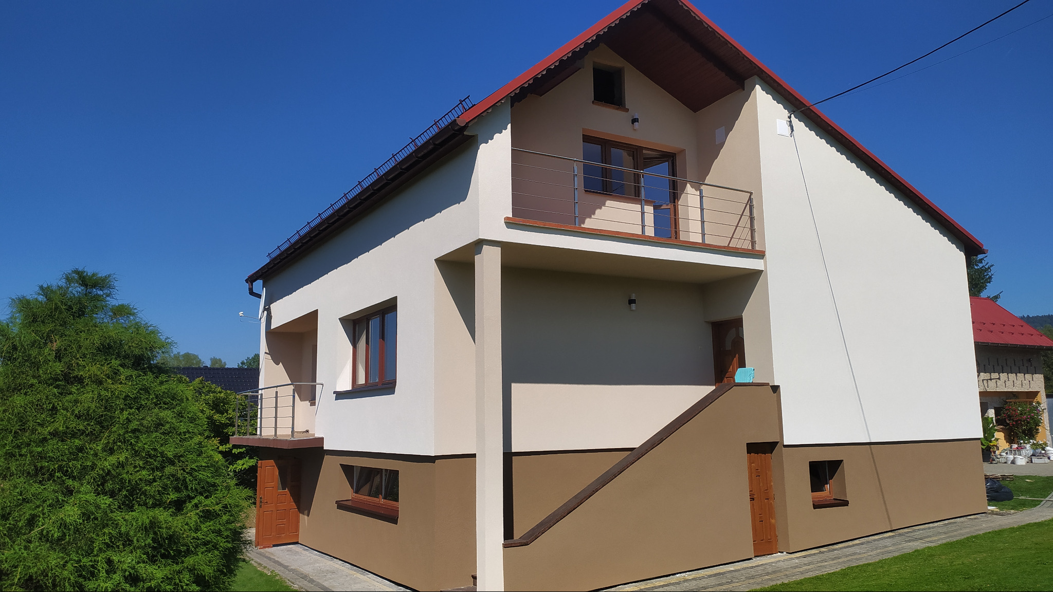 Dwukondygnacyjny dom jednorodzinny z balkonem, ocieplony i pomalowany w dwóch odcieniach beżu, z czerwoną dachówką, na tle błękitnego nieba i zieleni.