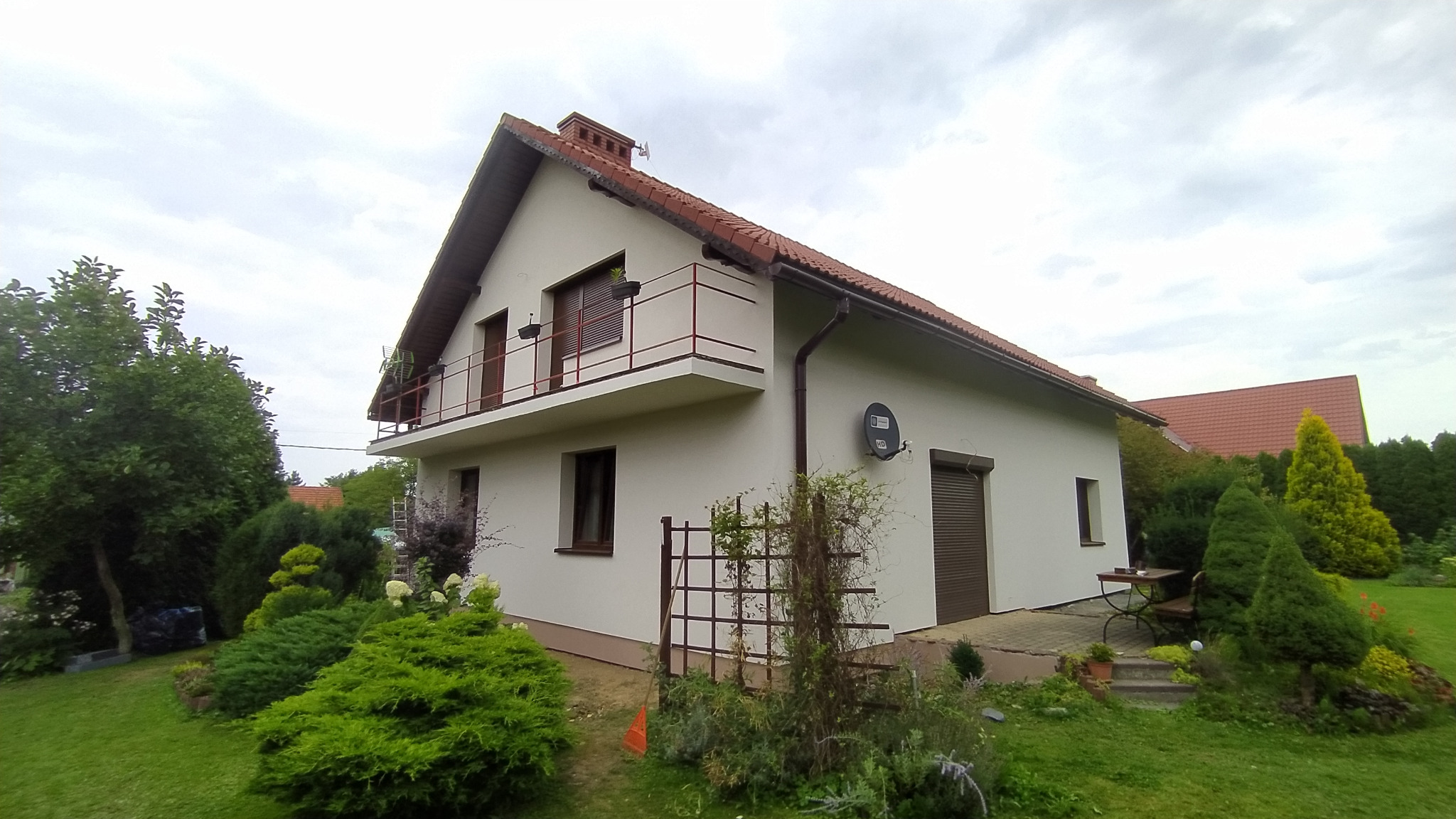 Dwupiętrowy dom z balkonem po renowacji elewacji, widoczny satelita na ścianie, brązowe okna i rolety, zadbany ogród z licznymi krzewami i drzewami.