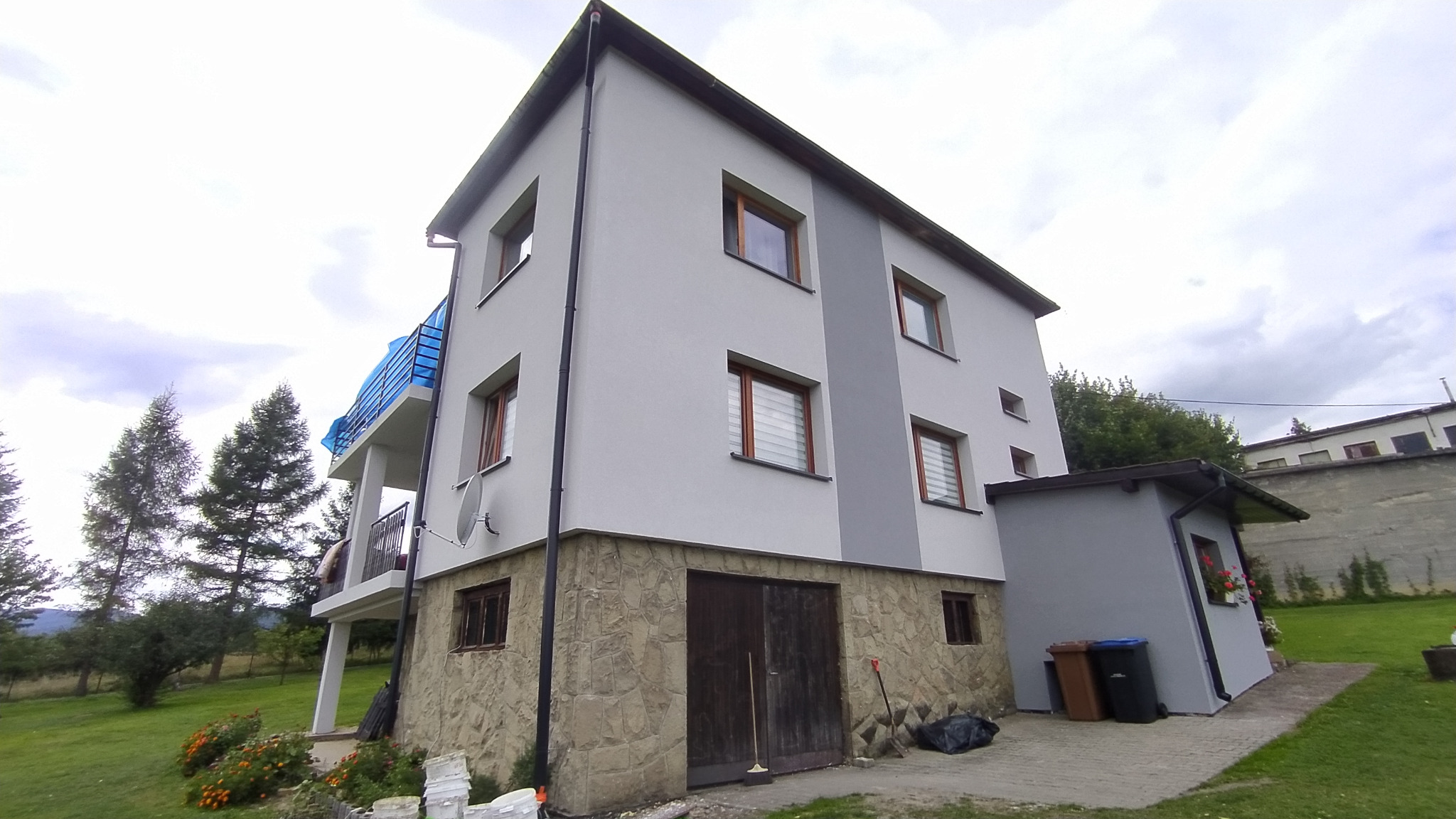 Dwukondygnacyjny dom z szarą elewacją i kamienną podmurówką, widok z niskiego kąta, z boku przylega szary budynek gospodarczy, na balkonie niebieska plandeka.