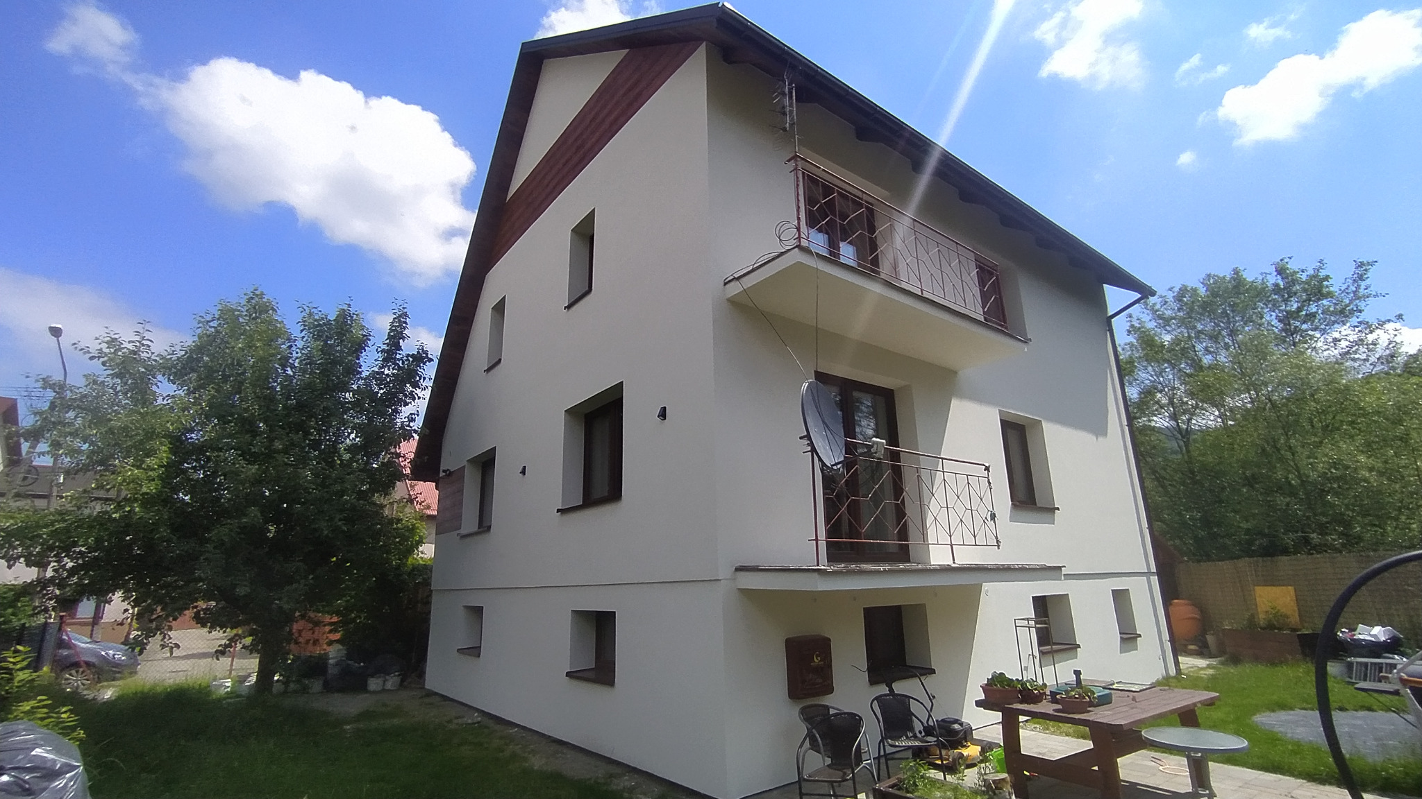 Dwupiętrowy dom z jasną elewacją, balkonami z metalowymi balustradami, widoczne okna z brązowymi ramami, antena satelitarna na balkonie, trawnik przed domem, błękitne niebo z chmurami w tle.