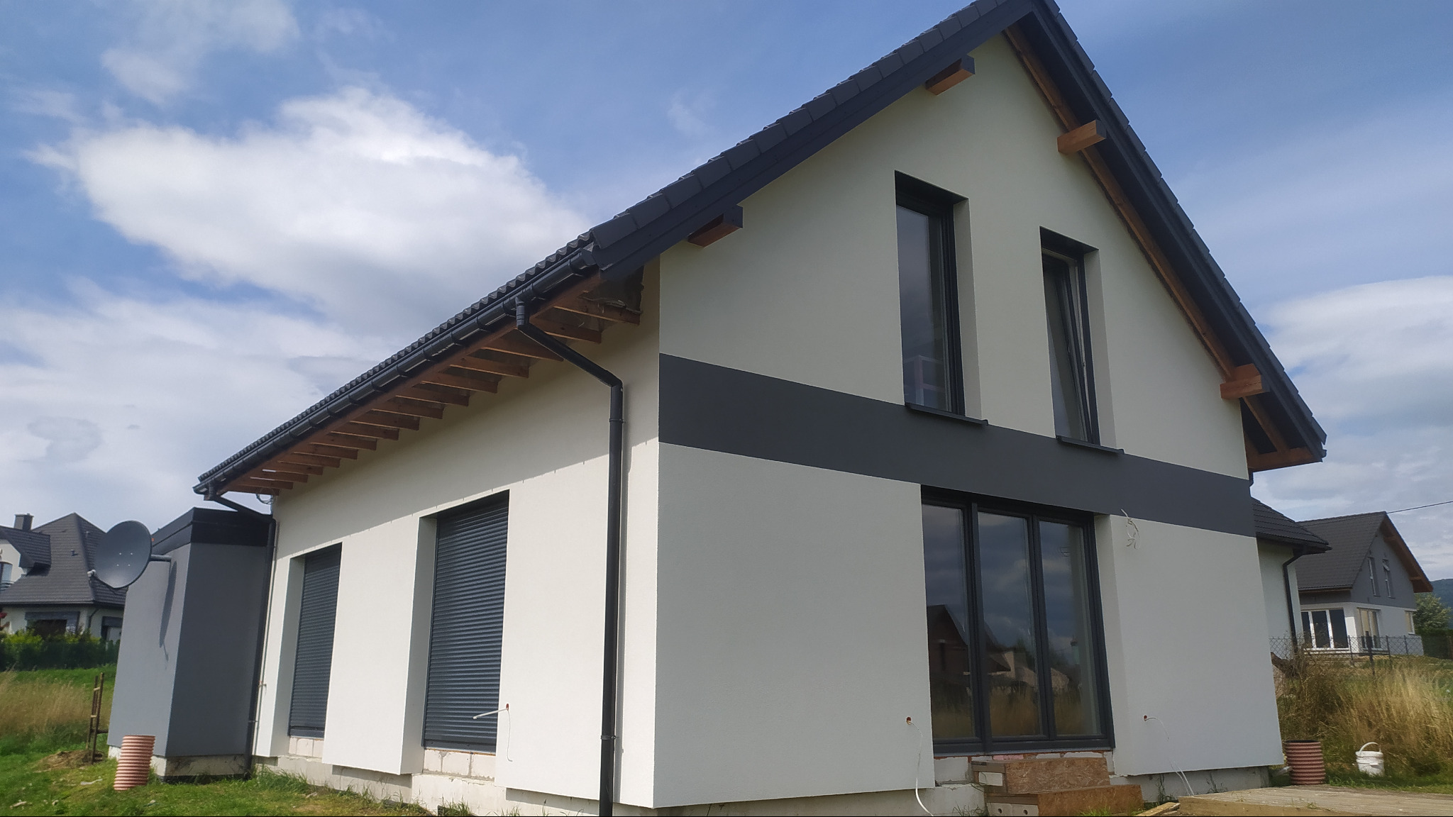 Nowoczesny dom jednorodzinny z jasną elewacją i ciemnoszarym pasem, widoczne okna z roletami, orynnowanie i fragment dachu, w tle błękitne niebo z chmurami oraz trawa.