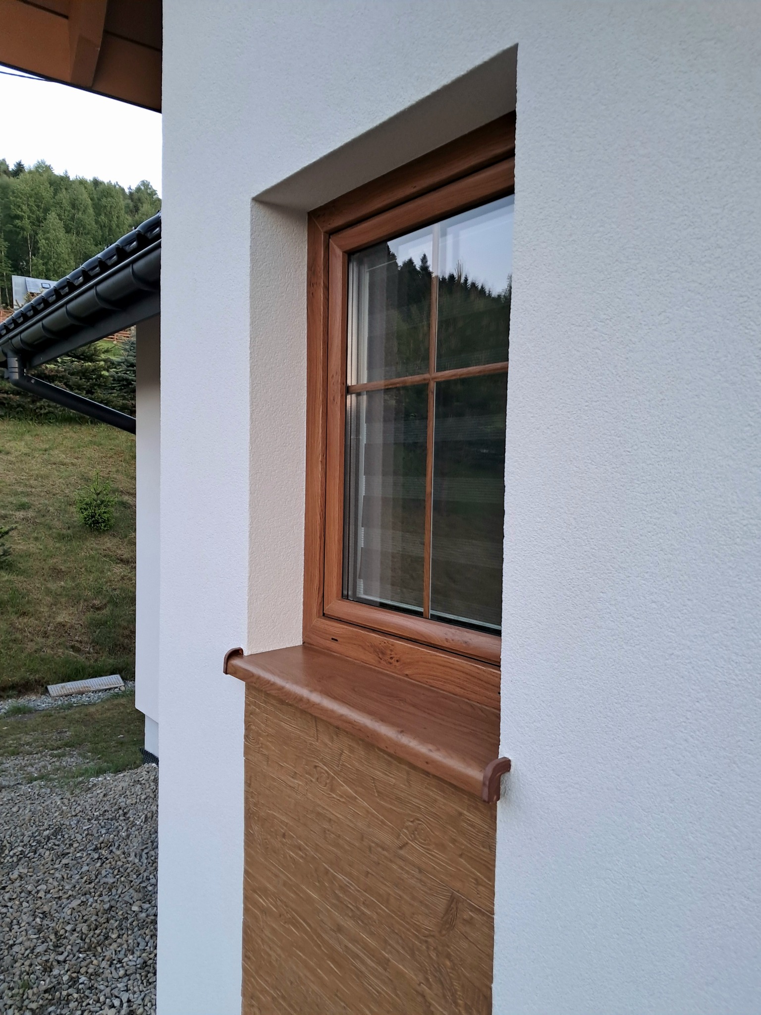 Detal wykończenia elewacji z oknem w drewnianej ramie. Biała, teksturowana ściana z elementami dekoracyjnymi w kolorze drewna. Widok na zieleń w tle.