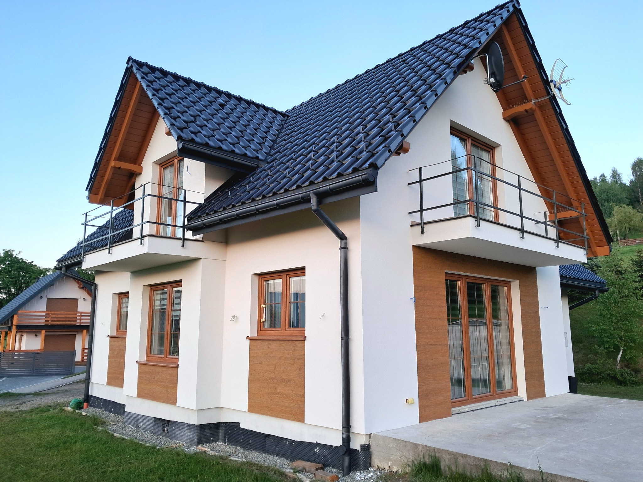 Nowoczesny dom z białą elewacją, elementami drewnopodobnymi i ciemną dachówką. Widoczne balkony z czarną balustradą i anteny na dachu. Dom po renowacji.