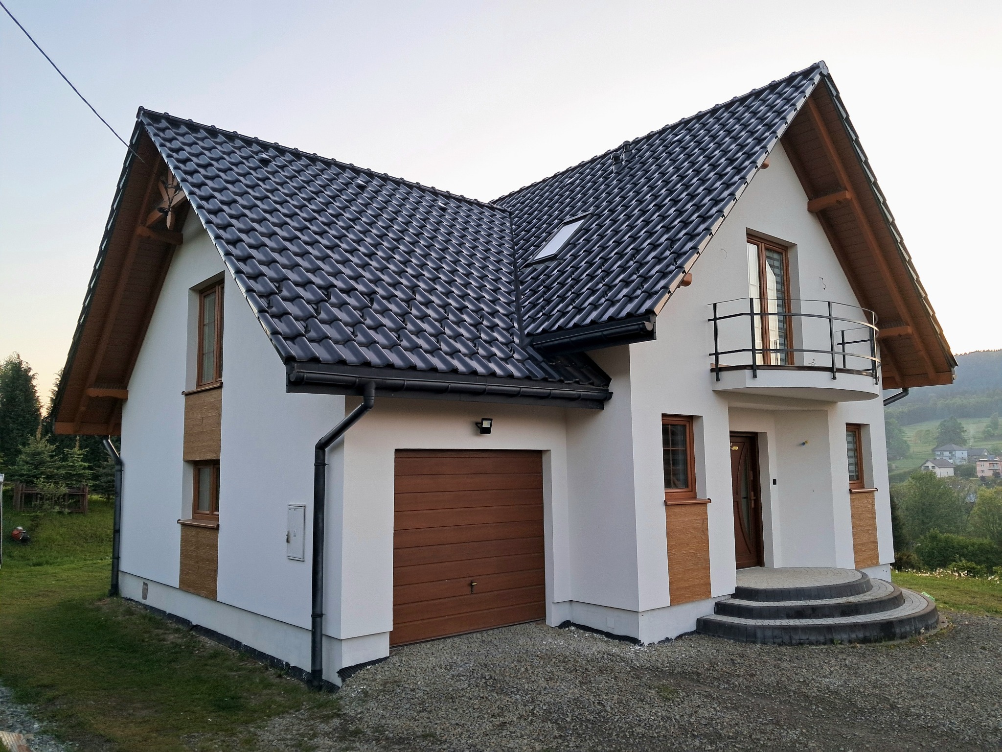 Nowoczesny dom jednorodzinny z białą elewacją, brązowymi akcentami i ciemną dachówką. Widoczny garaż, balkon i wejście z kamiennymi schodami.