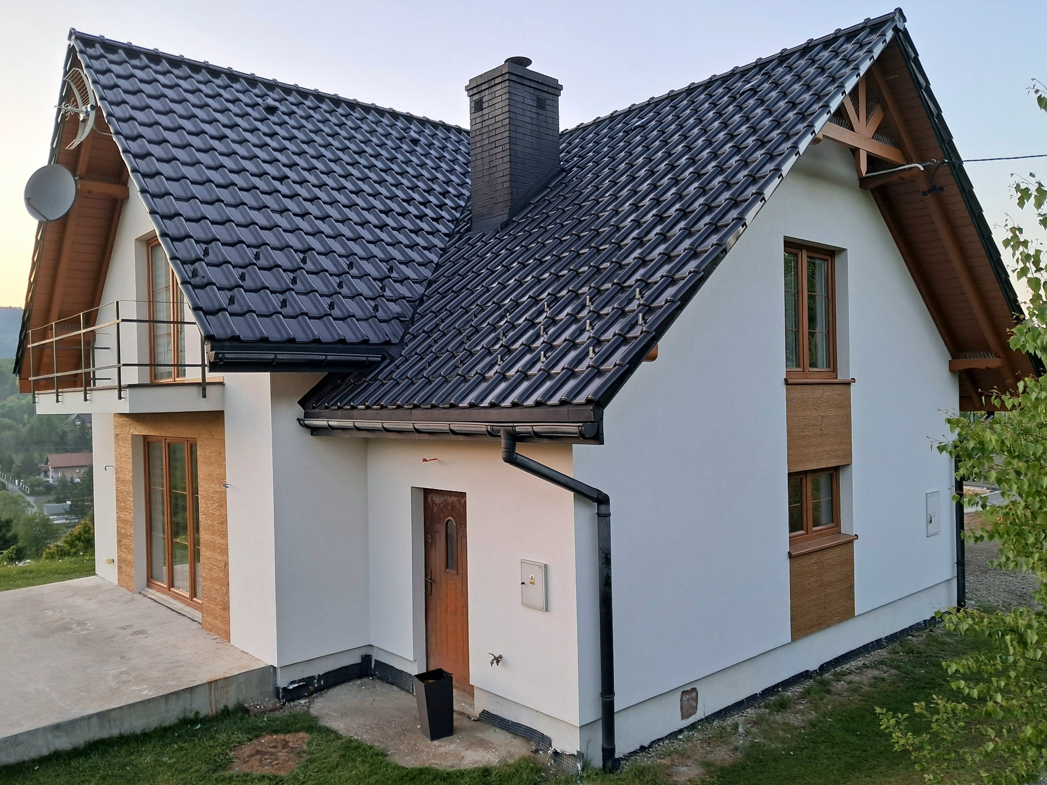 Nowoczesny dom jednorodzinny z białą elewacją, ciemną dachówką i drewnianymi akcentami. Widoczny komin, okna i drzwi wejściowe. Ujęcie z zewnątrz, w pogodny dzień.