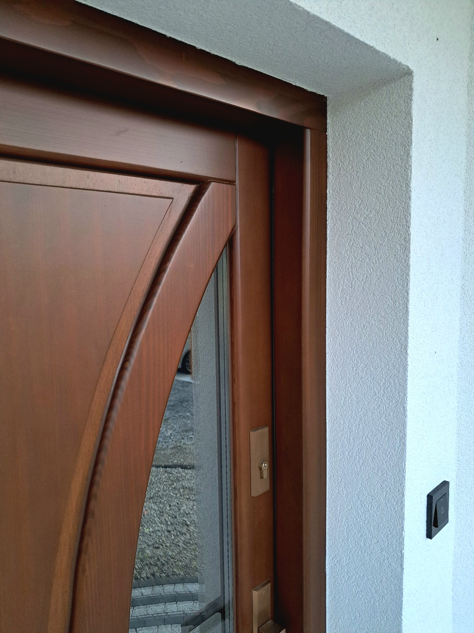 Wejście do domu: brązowe drzwi z szybą i biała, teksturowana elewacja z dzwonkiem. Detal wykończenia i stolarki.