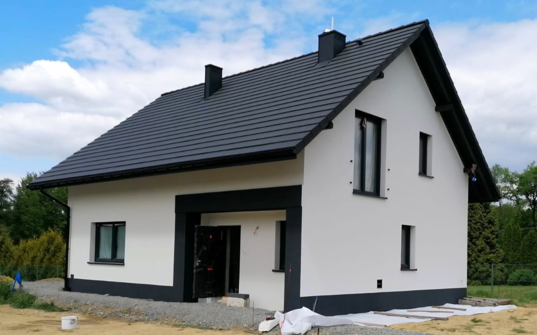 Nowoczesny dom jednorodzinny z białą elewacją i czarnymi akcentami, widok z zewnątrz, częściowo zabezpieczone wejście, teren wokół domu w trakcie prac wykończeniowych.