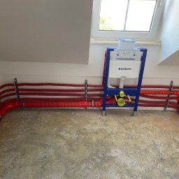 Ryszard Skowyrski - Instalacja podtynkowa toalety Geberit z czerwonymi rurami w izolacji termicznej, zamocowanymi do ściany na metalowych uchwytach, na surowej betonowej podłodze w pomieszczeniu ze skosami i oknem...