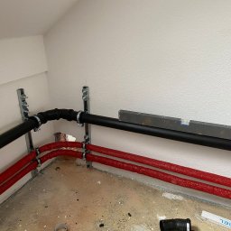 Ryszard Skowyrski - Nowe rury hydrauliczne w czerwonej izolacji oraz czarna rura ułożone równolegle przy ścianie w trakcie montażu w Krakowie, poziomica na rurze.