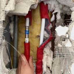 Ryszard Skowyrski - Instalacja hydrauliczna w trakcie naprawy: białe rury z czerwoną izolacją, metalowe złączki, widoczne warstwy izolacji ściany i instalacji, ręka montera przytrzymująca rurę.