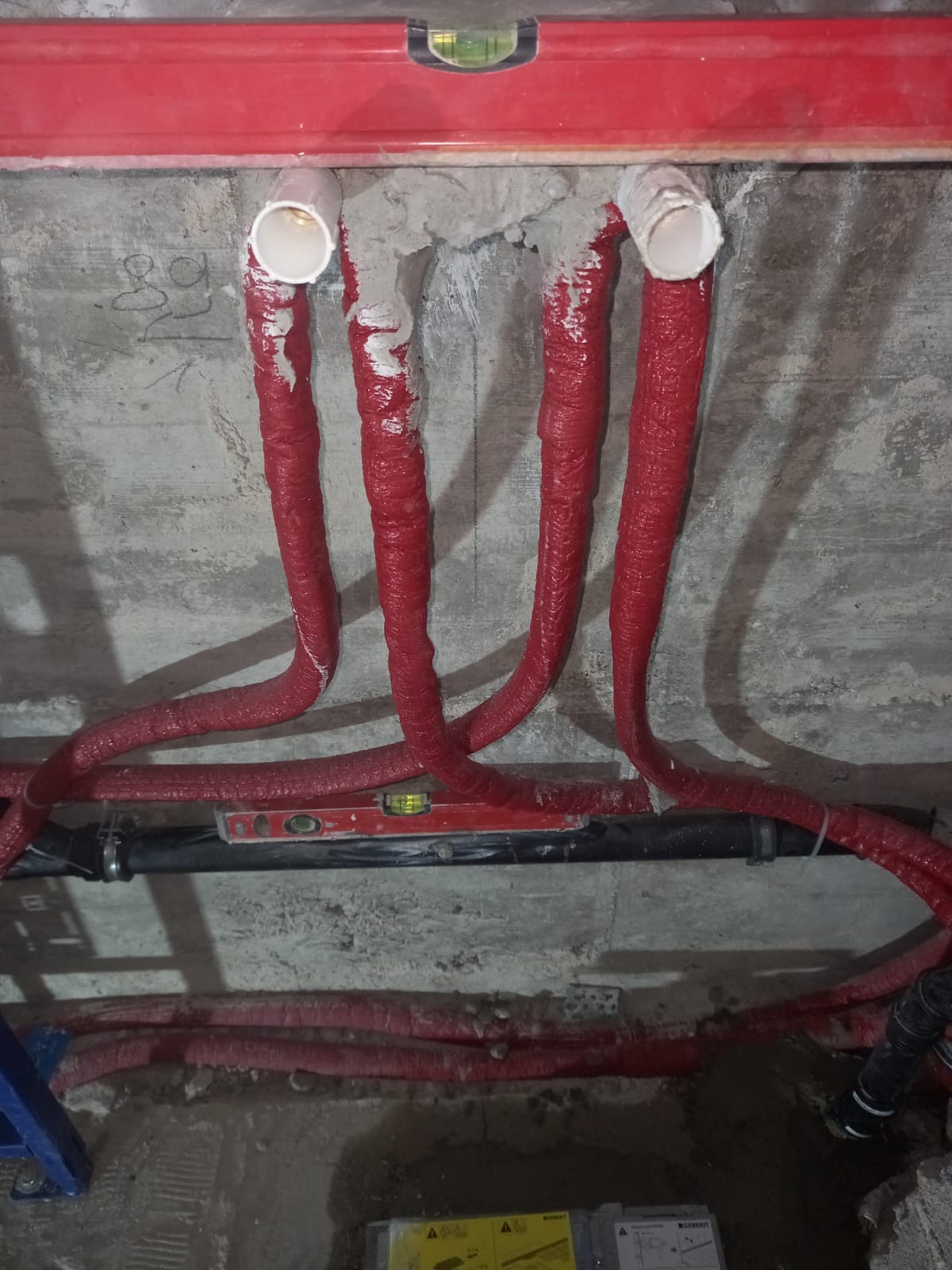 Czerwone, zaizolowane rury hydrauliczne wystające ze ściany, zabezpieczone zaprawą, z widoczną poziomicą czerwoną i czarną rurą w tle.