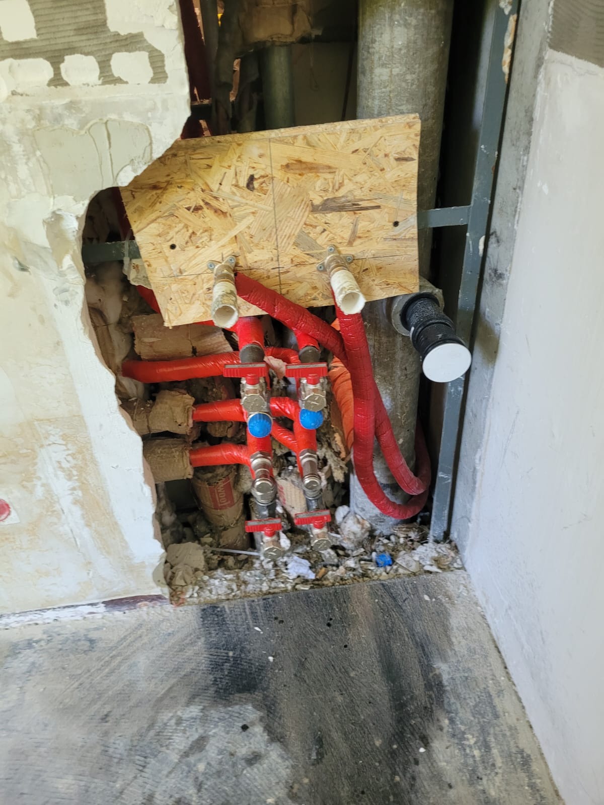 Fragment instalacji hydraulicznej z czerwonymi rurami i zaworami z niebieskimi pokrętłami, zamocowanej do płyty OSB w ścianie z widocznym ubytkiem tynku.