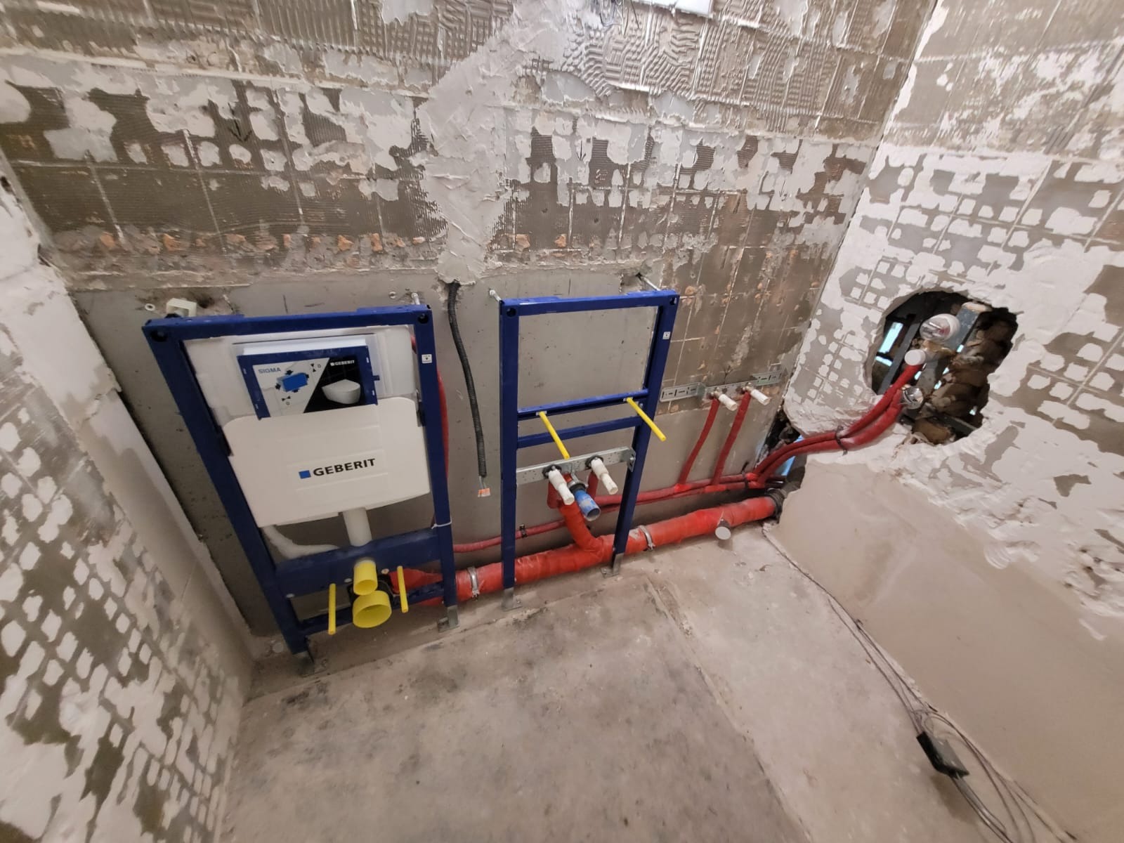 Montaż stelaża podtynkowego Geberit z instalacją hydrauliczną w surowym wnętrzu łazienki, widoczne rury w kolorze czerwonym i żółtym na tle otynkowanej ściany.