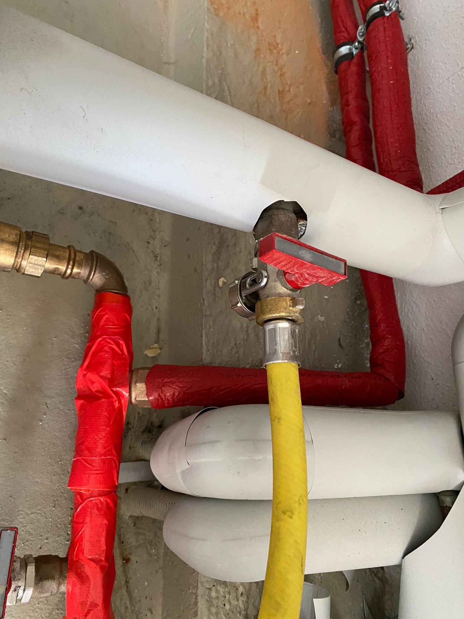 Fragment instalacji hydraulicznej z zaworem z czerwoną rączką, żółtym wężem i rurami w izolacji termicznej, widoczne elementy miedziane.