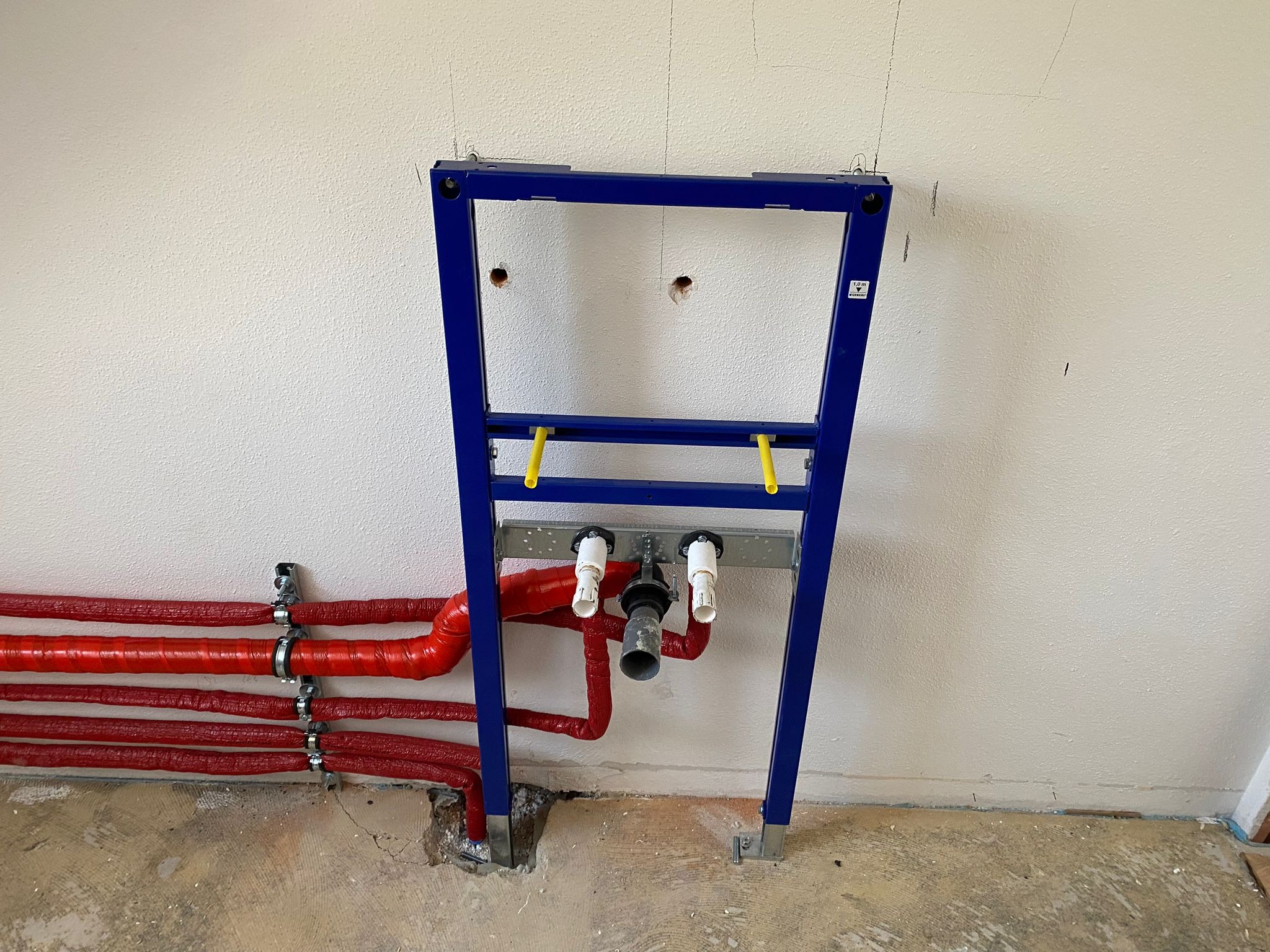 Stelaż podtynkowy WC w trakcie montażu, widoczne czerwone rury instalacji wodnej w izolacji, surowa ściana i podłoga w trakcie remontu.