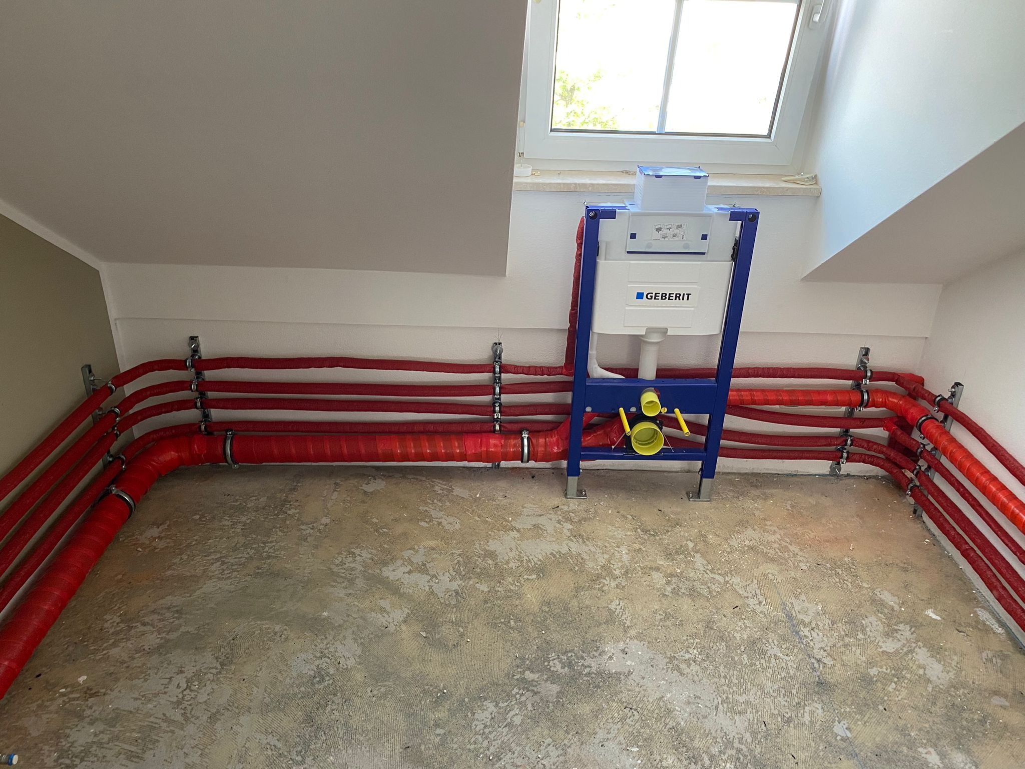 Instalacja podtynkowa toalety Geberit z czerwonymi rurami w izolacji termicznej, zamocowanymi do ściany na metalowych uchwytach, na surowej betonowej podłodze w pomieszczeniu ze skosami i oknem...