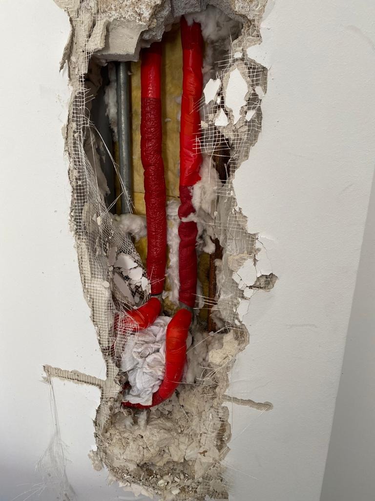 Odsłonięte, czerwone rury instalacji wodnej w ścianie, otoczone izolacją i fragmentami gruzu po kuciu.