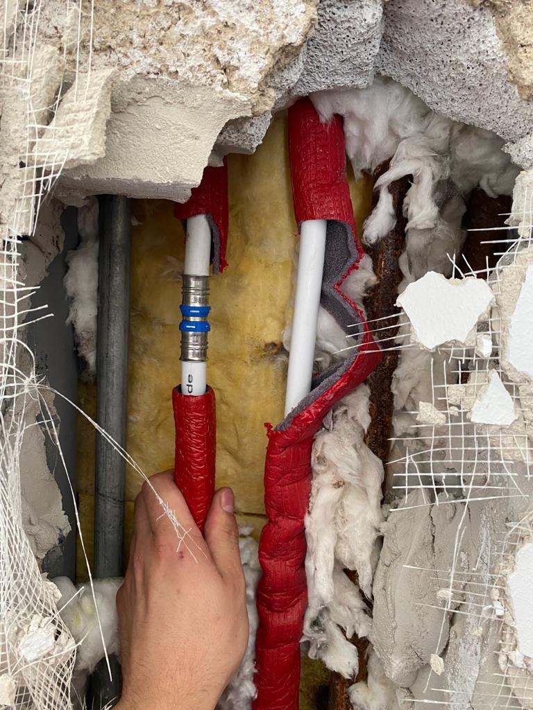Instalacja hydrauliczna w trakcie naprawy: białe rury z czerwoną izolacją, metalowe złączki, widoczne warstwy izolacji ściany i instalacji, ręka montera przytrzymująca rurę.