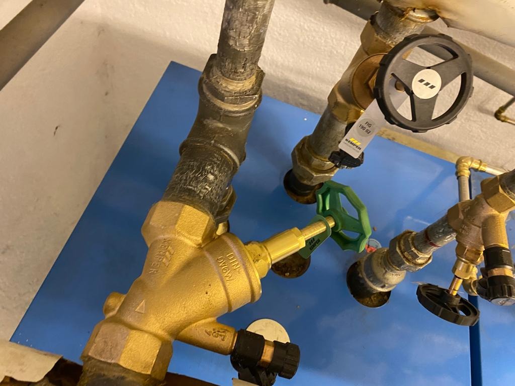 Złożony układ rur hydraulicznych z zaworami Kemper, widoczne różne typy połączeń i materiałów, tło stanowi niebieska płyta montażowa.