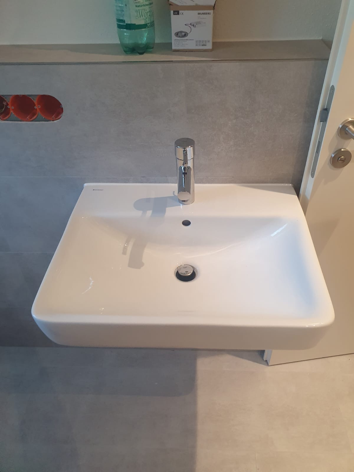 Biała, prostokątna umywalka z chromowaną baterią, zamontowana na szarej ścianie z butelką wody i pudełkiem w tle.