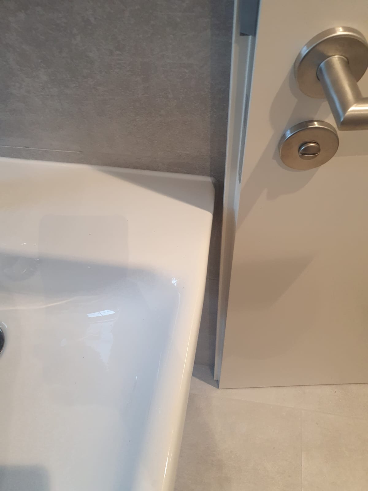 Fragment umywalki ceramicznej z widocznym odpływem, szara ściana z fakturą betonu oraz fragment białych drzwi z metalową klamką.