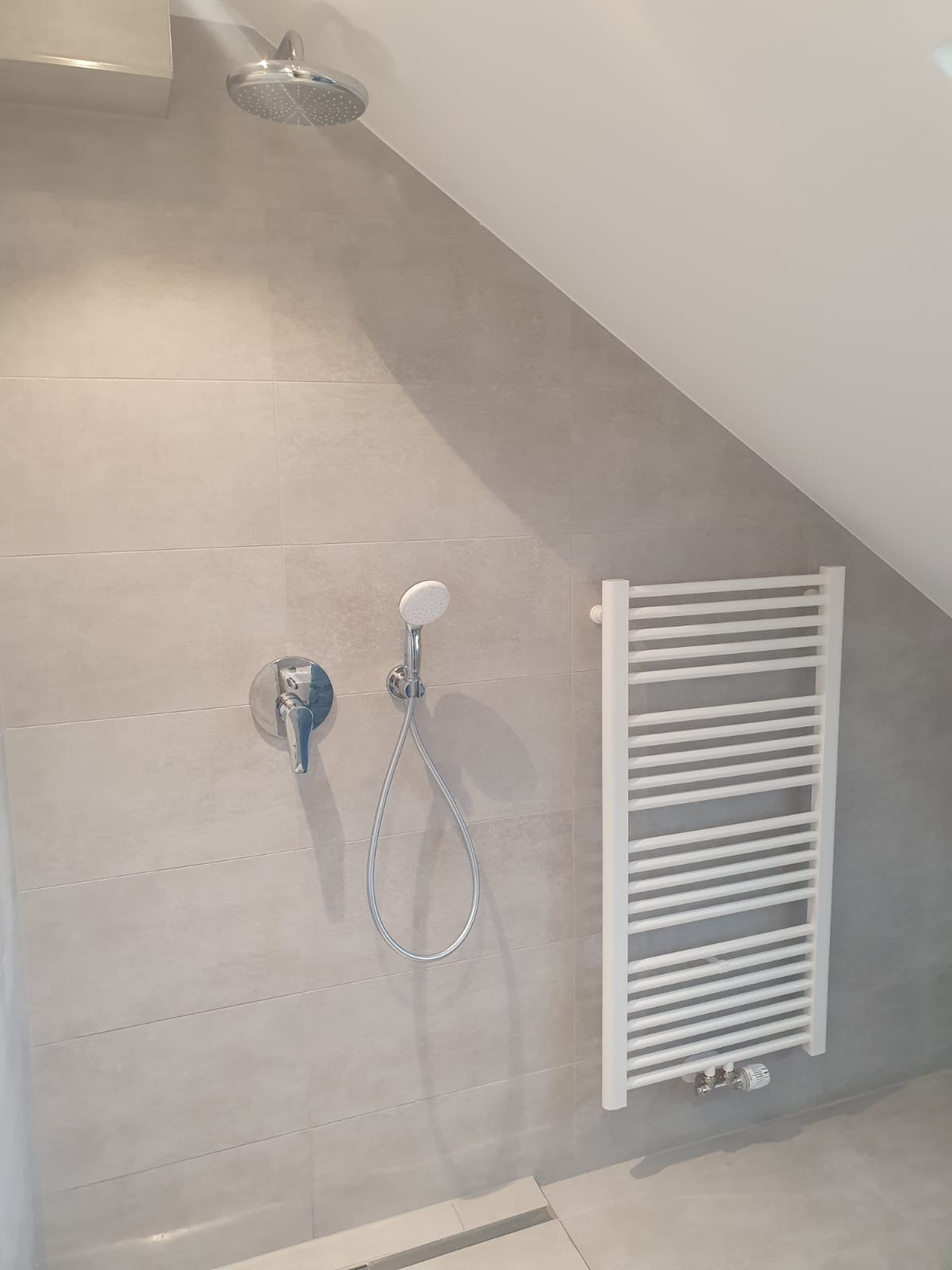 Wykończona łazienka na poddaszu ze srebrną armaturą prysznicową i białym grzejnikiem drabinkowym, szare płytki ścienne i podłogowe, widoczny odpływ liniowy.