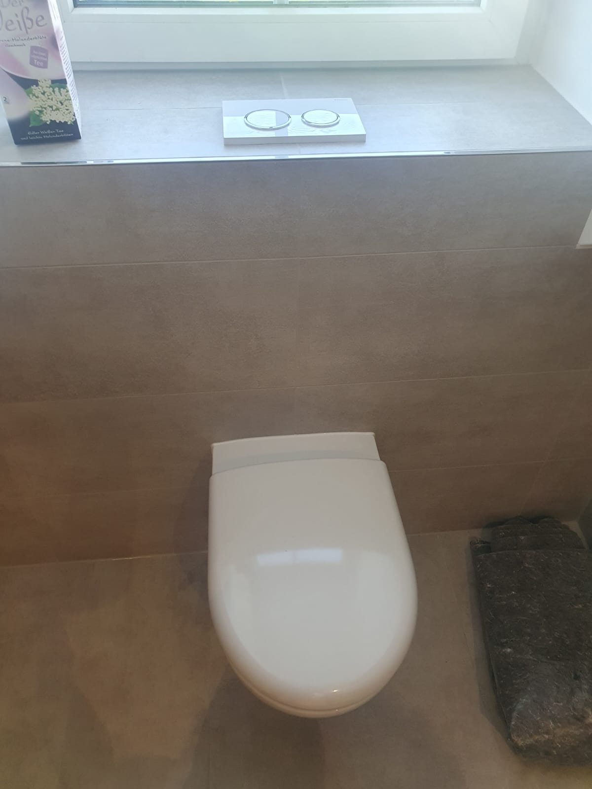 Zamontowana podwieszana toaleta z przyciskiem spłukującym dwufunkcyjnym i dekoracyjnym kamieniem obok, na tle płytek ceramicznych.