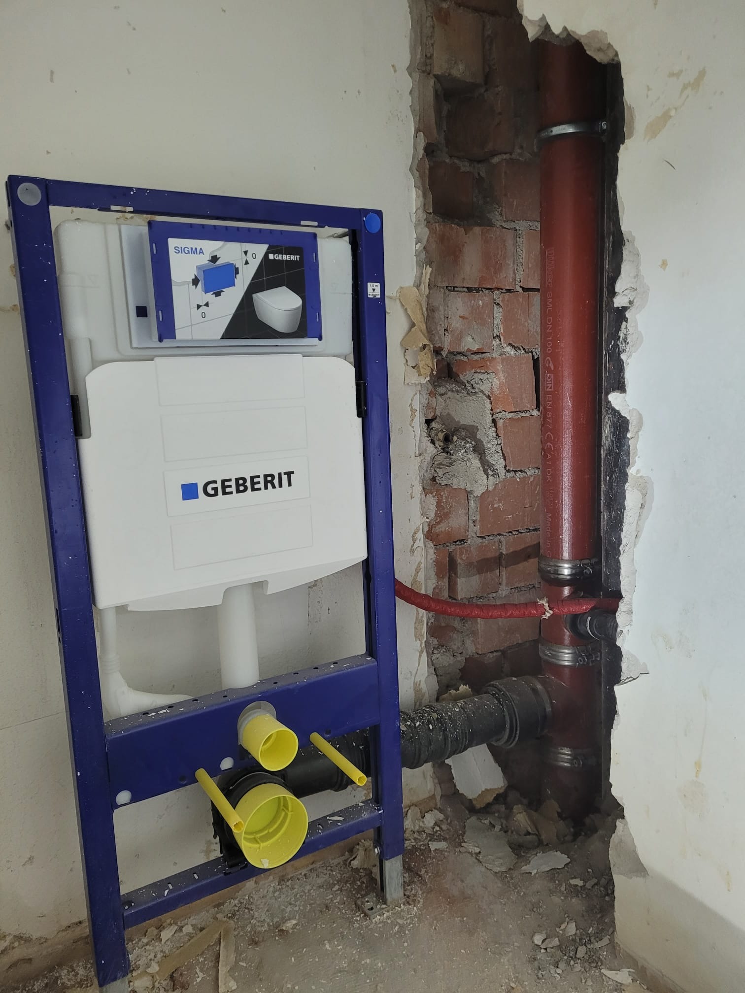 Stelaż podtynkowy Geberit Sigma z widocznymi przyłączami kanalizacyjnymi i rurą odpływową w surowej ścianie z cegły, przed zabudową.